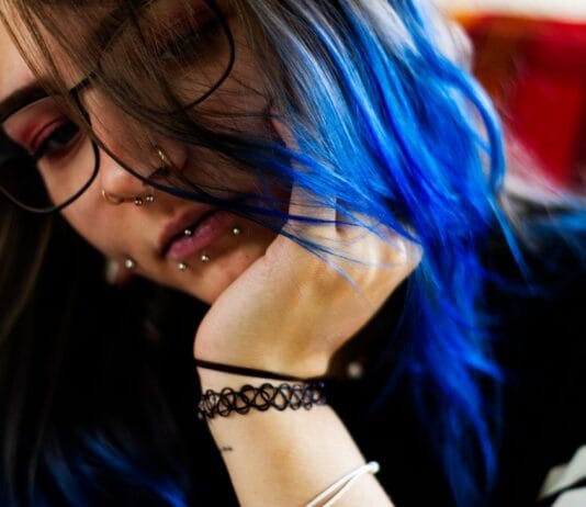 Czy piercing w brwi zostawia ślad na zawsze? Pytania czytelników o blizny po kolczykach Młoda kobieta z niebieskimi włosami, w okularach i z kilkoma piercingami